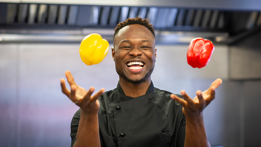 A Sanctuary Care chef wearing chef blacks juggling with a yellow and red pepper whilst laughing