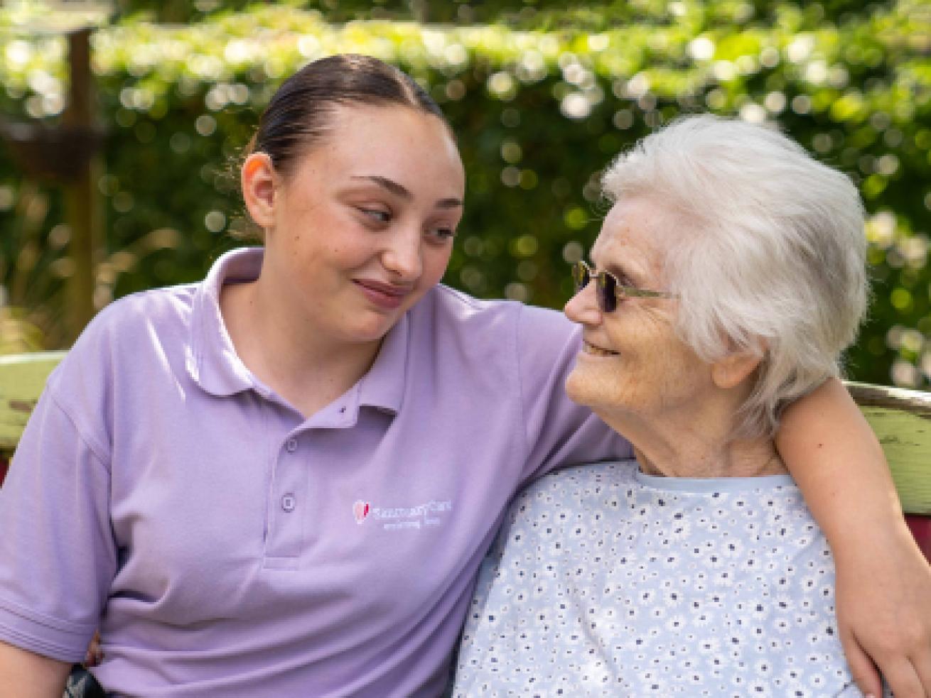 Sanctuary Care Assistant sitting on a garden bench with their arm around a resident